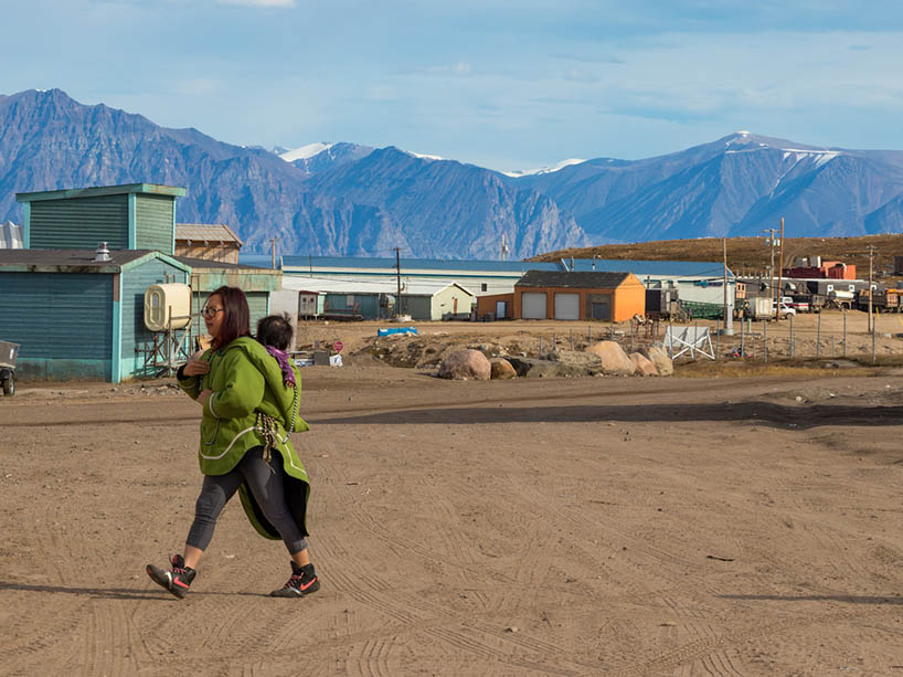Woman carrying baby on her back in a remote town with mountains in the background.
