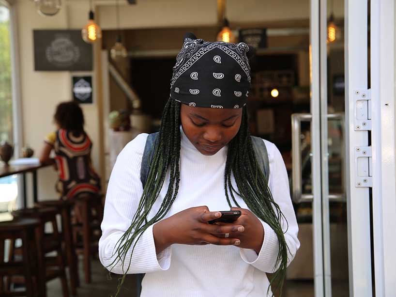 A young Black woman looks down at her phone while standing in a coffee shop. 