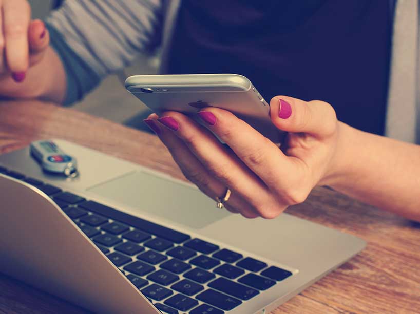 A woman holds a cell phone over a laptop.