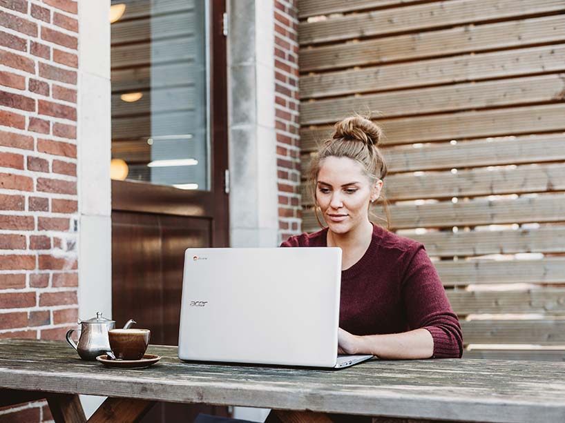 A young woman works on her computer sitting outside at a backyard table.
