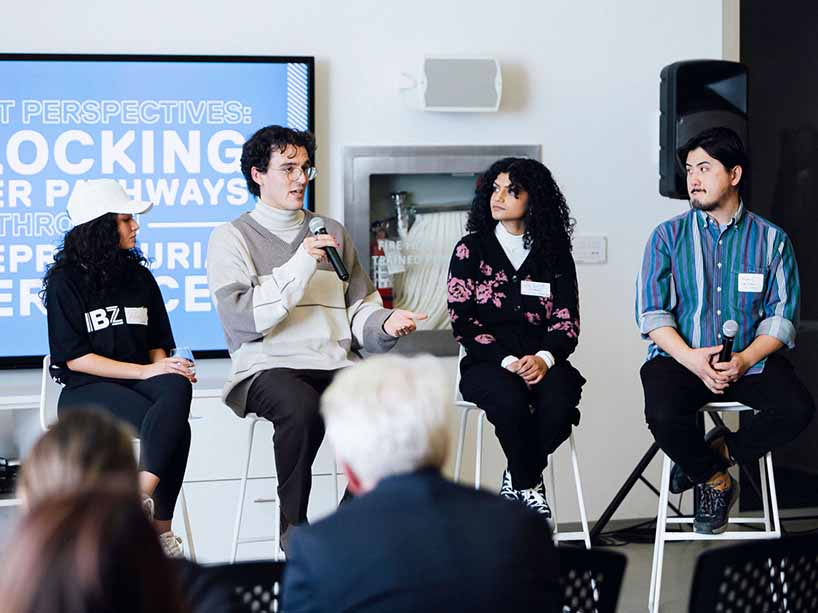 Four people sit on stools as part of a panel discussion, with one person speaking into a microphone.