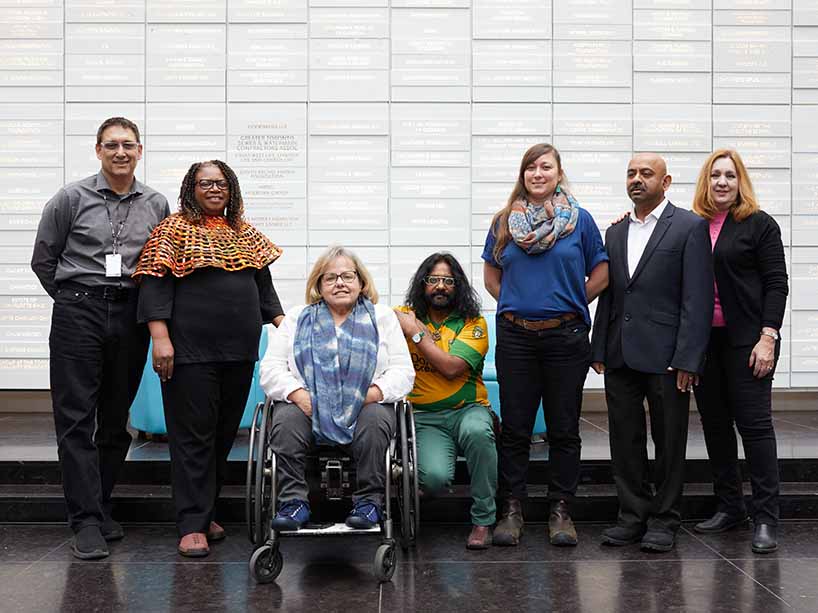 A group of seven people who are racially diverse, the woman in the centre is on a wheelchair and a man kneels next to her.