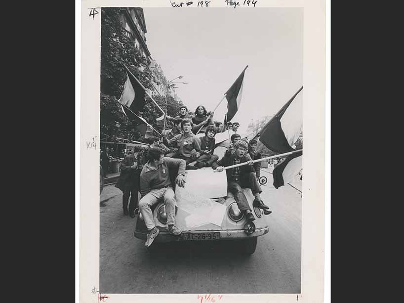A black and white photo of many young people piled onto a car, carrying flags