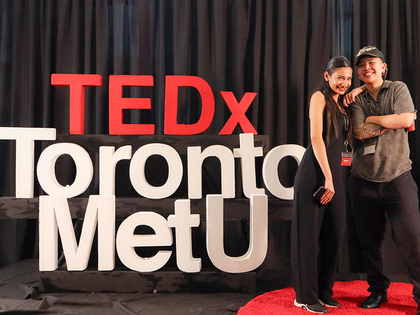 Two smiling students stand together beside a large “TEDxTorontoMetU” sign.