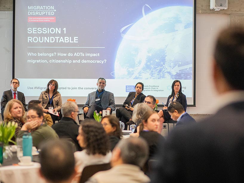  A room full of people facing a panel of speakers with a projector screen behind them.