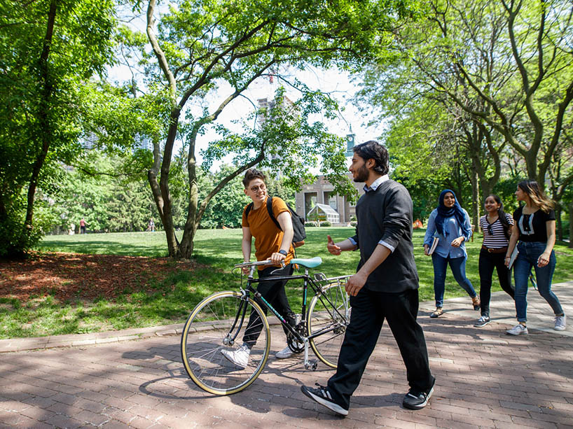 Two people walking on campus, one of them holds a cycle.