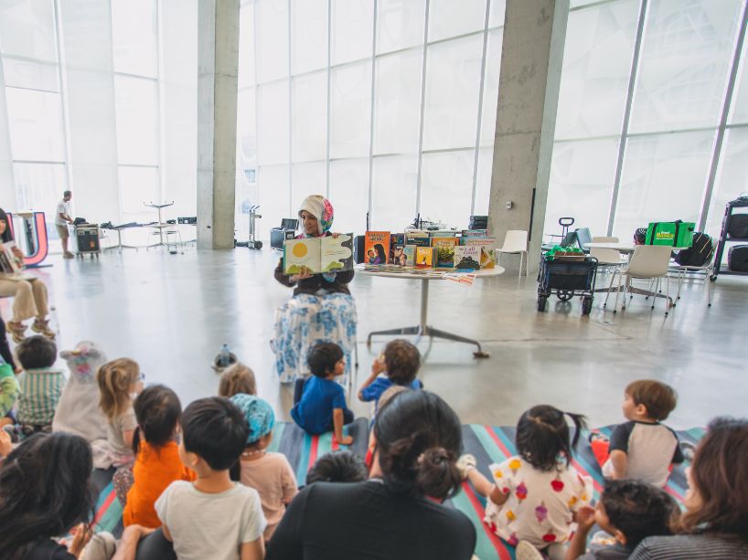 A group of children sit on the floor together as someone on a chair reads them a story.
