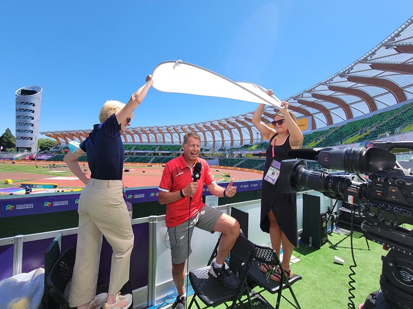 A journalist stands in a stadium with a microphone and two staff hold a round cover over him.