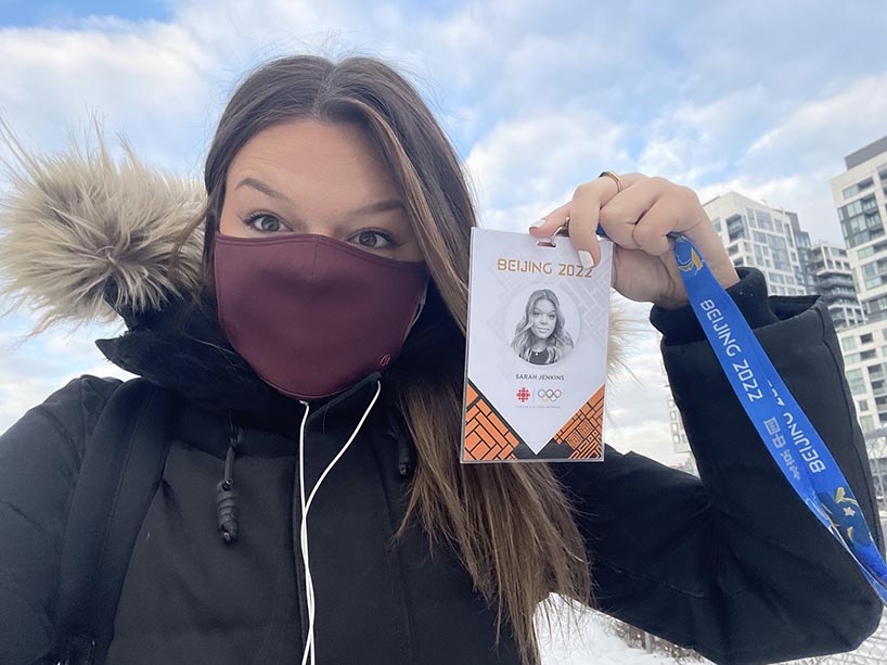 Sarah Jenkins in a winter coat and mask, holding up her Beijing 2022 Olympic name badge pass from when she worked the event for CBC.