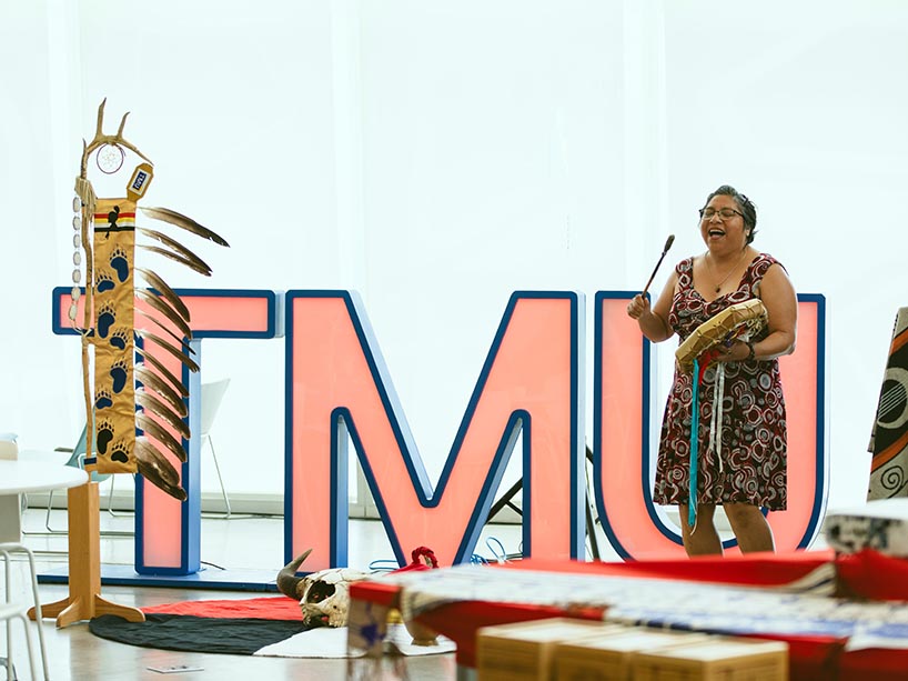 Amy Desjarlais sings and plays the hand drum, standing in front of large “TMU” letters and the TMU eagle staff.