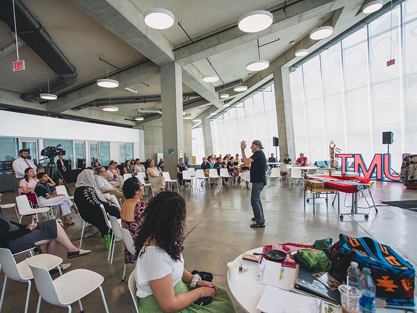 A room full of people sitting on white chairs in a circle, watching a presentation.