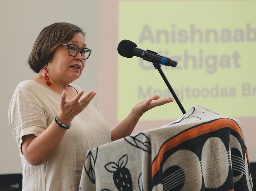 Monica McKay speaks into a microphone with her palms open to the sky.
