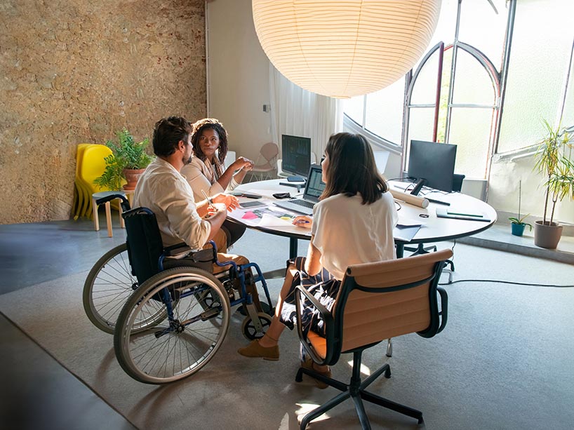 Three people talking and working at a table. Two people are in office chairs, and one is in a wheelchair.