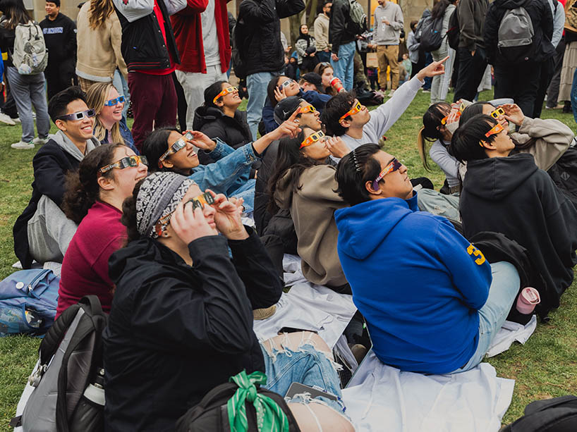 A crowd seated in the quad, wearing eclipse glasses, with some people pointing towards the sky.