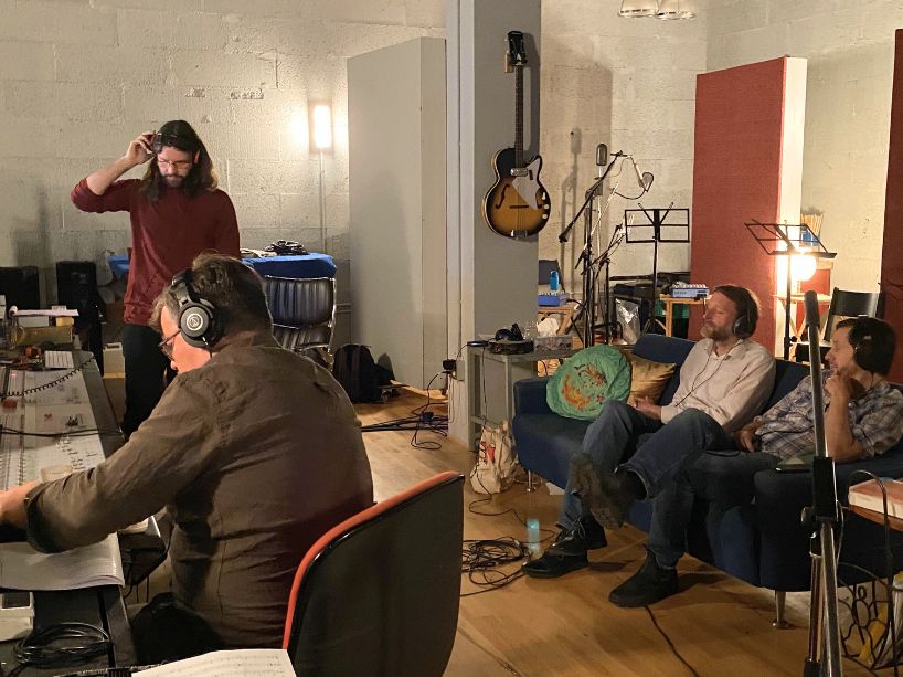 Band members sitting in the studio with instruments and production equipment. 
