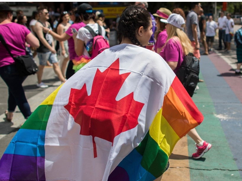 Young woman participating in the Pride Parade wears a Canada flag with rainbow edging.