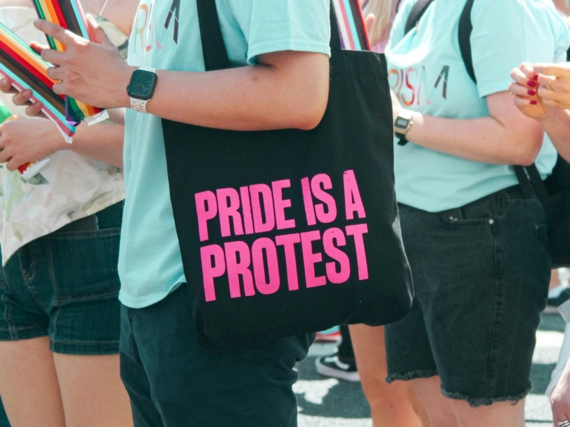 A black tote bag worn by a person reading ‘Pride is a protest’ in neon pink.