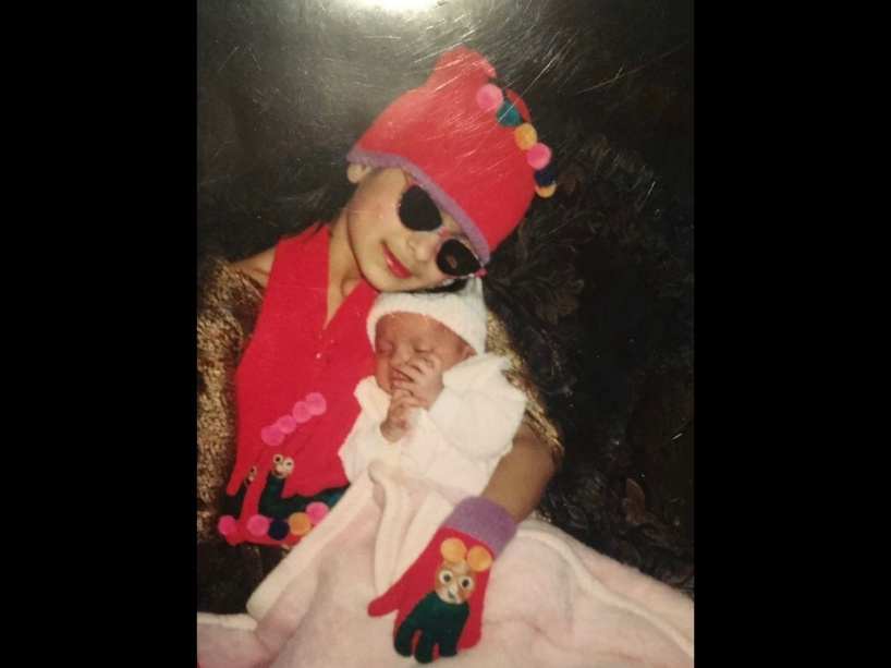 A young Sonia, wearing a red vest and hat, holds her baby sister Shabnam, who is dressed in a white outfit.