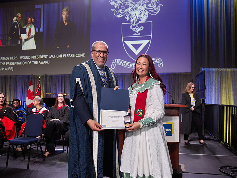 Two people pose for a photo at a ceremony while holding a certificate and a medal.
