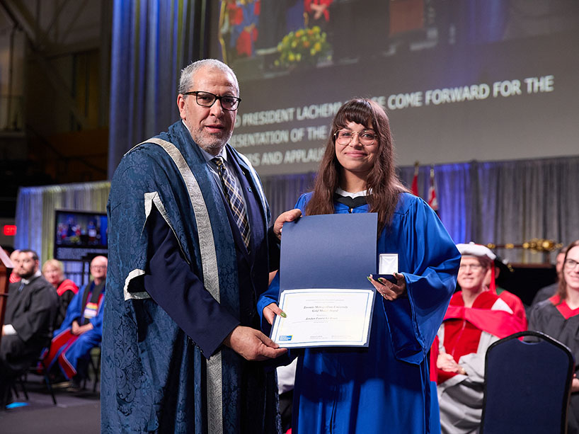 Two people wearing academic regalia pose for a photo while one holds a certificate and a medal.