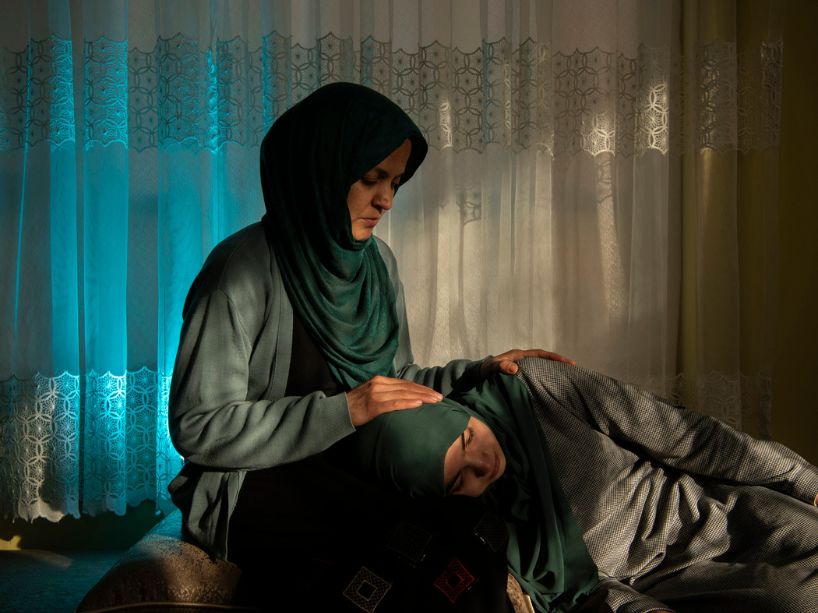 A woman in a green hijab gently cradles her daughter’s head in her lap, offering comfort.