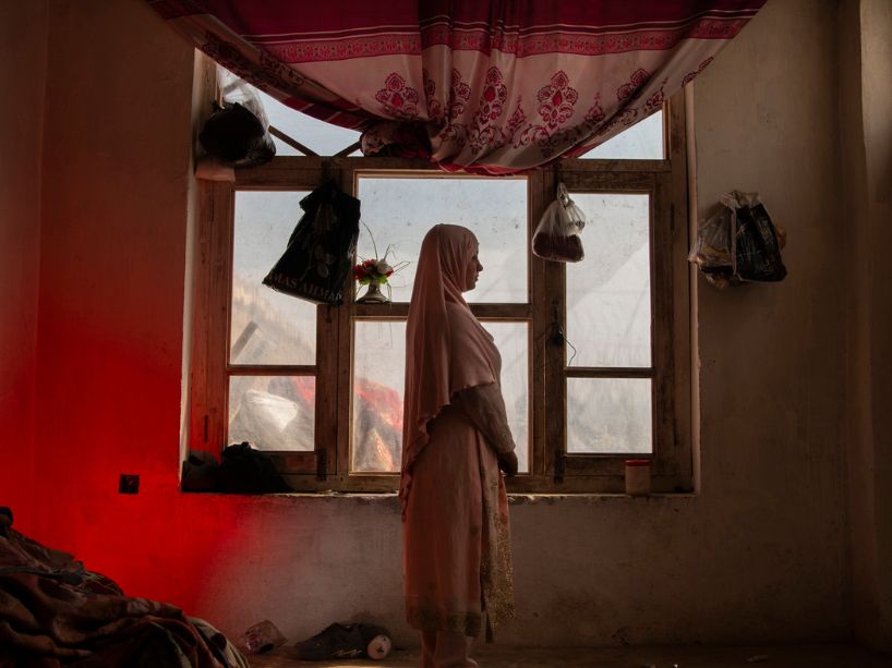 A young woman wearing a hijab stands in front of a window, quietly looking out into the distance. 