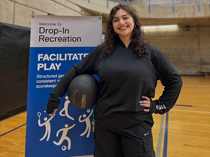 A female university student poses in a gym with a basketball under her arm.