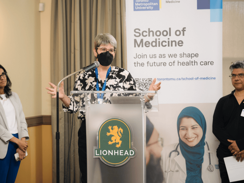  A person with a mask on speaking at a podium, flanked by two people.