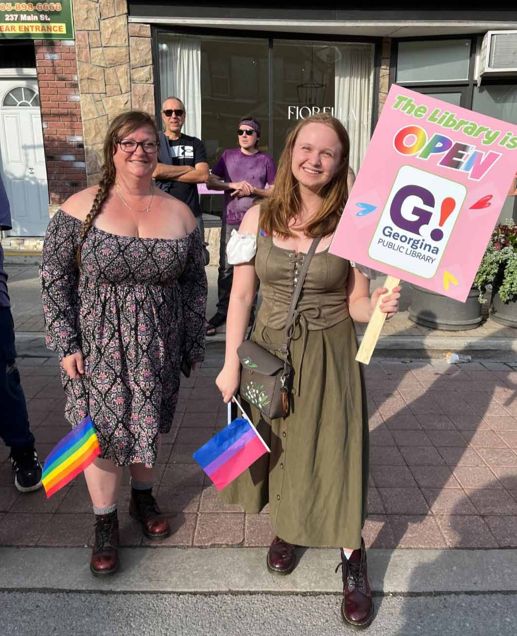 Two people post for a photo a Pride parade. One person is holding a sign that says “The Library is Open, Georgina Public Library”