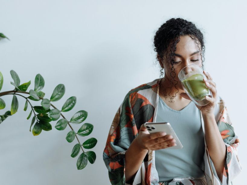 Young woman using a cellphone while drinking a glass of green matcha.
