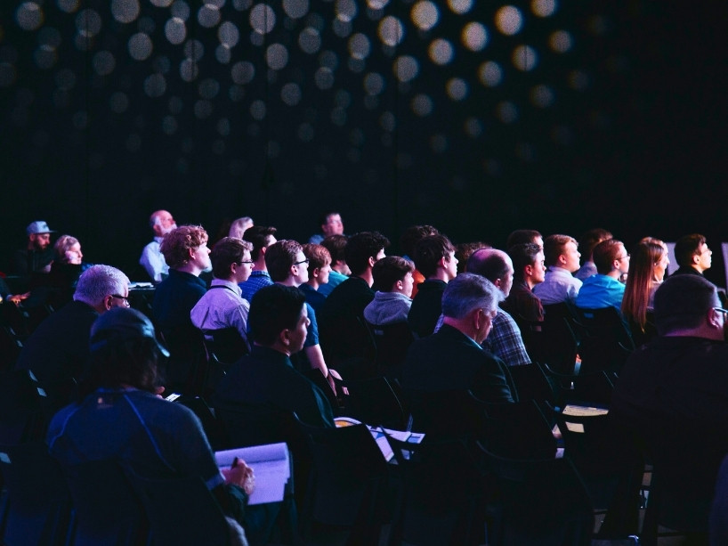 Audience members sit in a darkened auditorium looking toward a stage.
