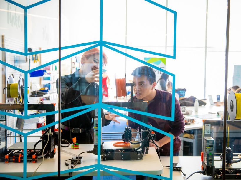 Two students work on a 3D printing project together using a 3D printer in a lab, in front of a teal coloured modern divider.