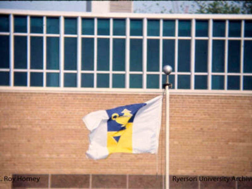 White, blue and yellow flag flies in the wind against a brick building with windows.