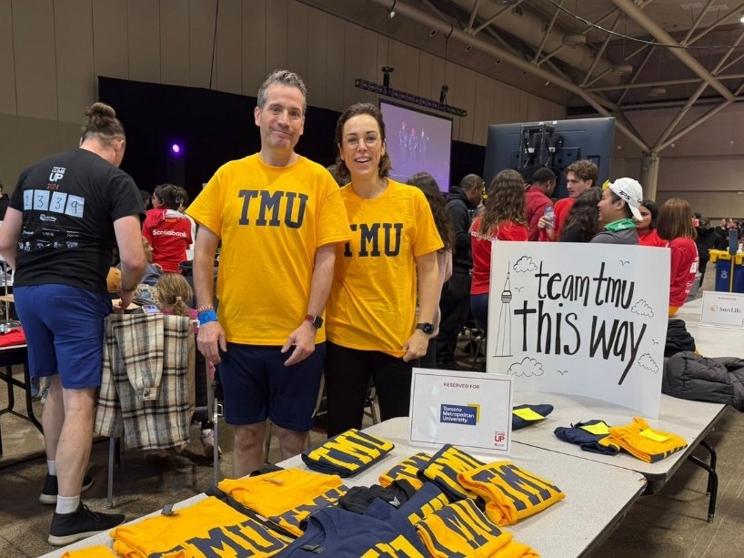 Two TMU community members wait at a table with TMU shirts.