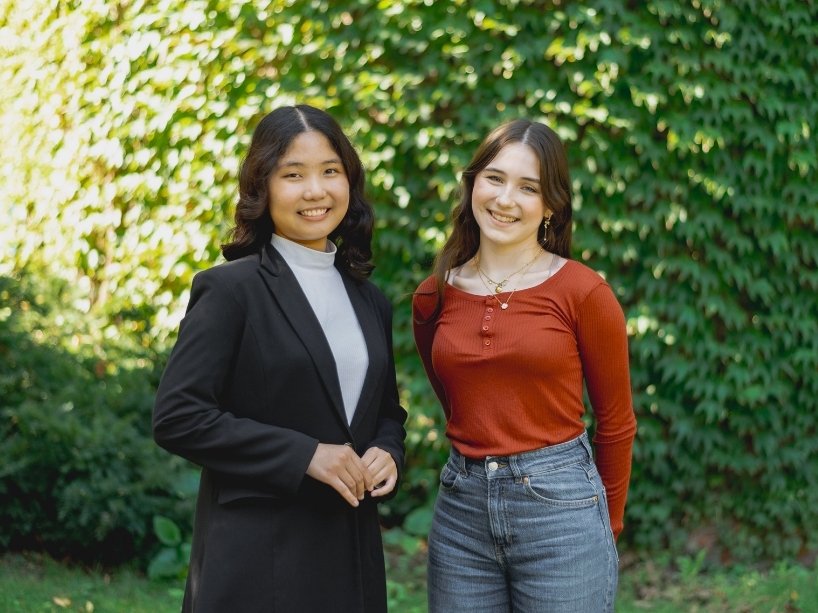 Two young women standing outside against a backdrop of bushes.