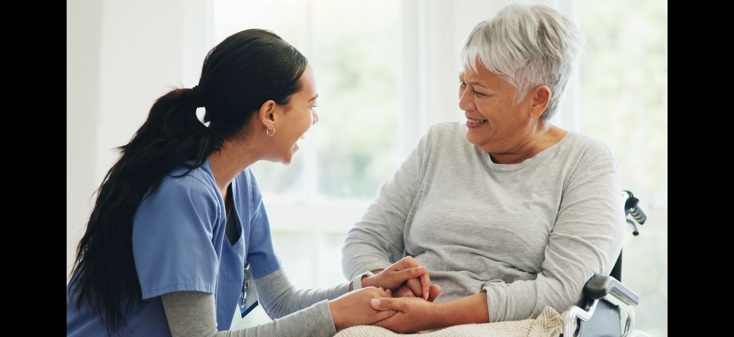 A woman in medical scrubs holds hands with an older woman in a wheelchair as they sit in the living room of a home.