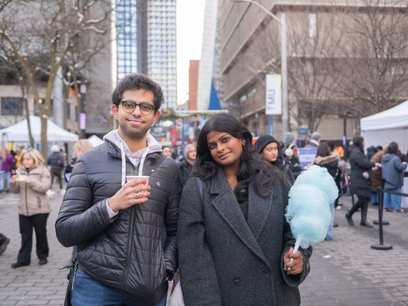 Two people pose for a photo outside while holding a hot beverage and blue cotton candy.