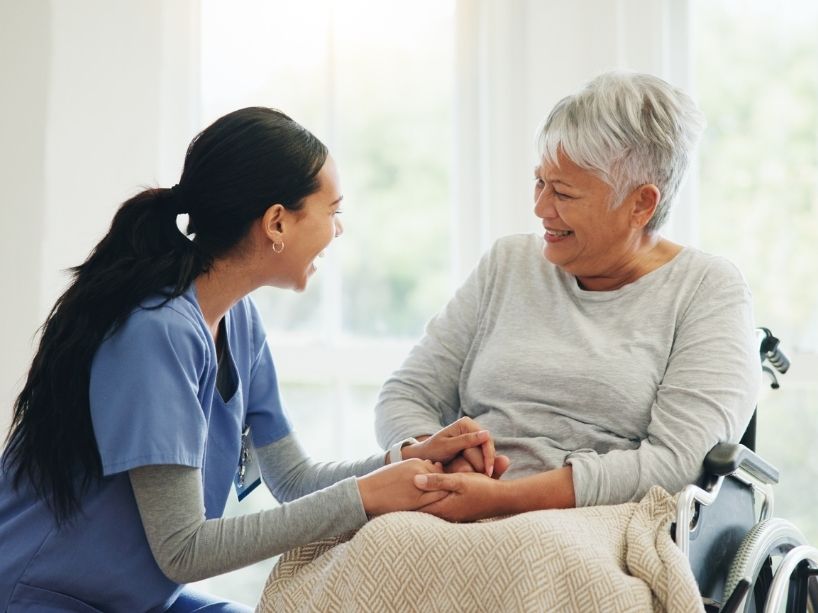 A woman in medical scrubs holds hands with an older woman in a wheelchair as they sit in the living room of a home.