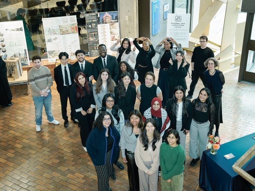 A group of university students and a professor pose for a photo at a design exhibit.