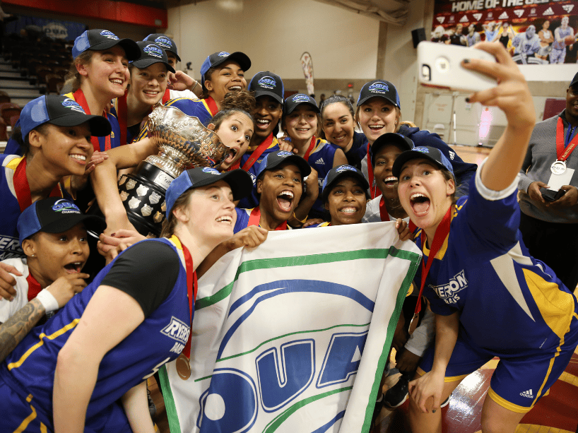 Women's basketball team taking a selfie with a trophy and banner.