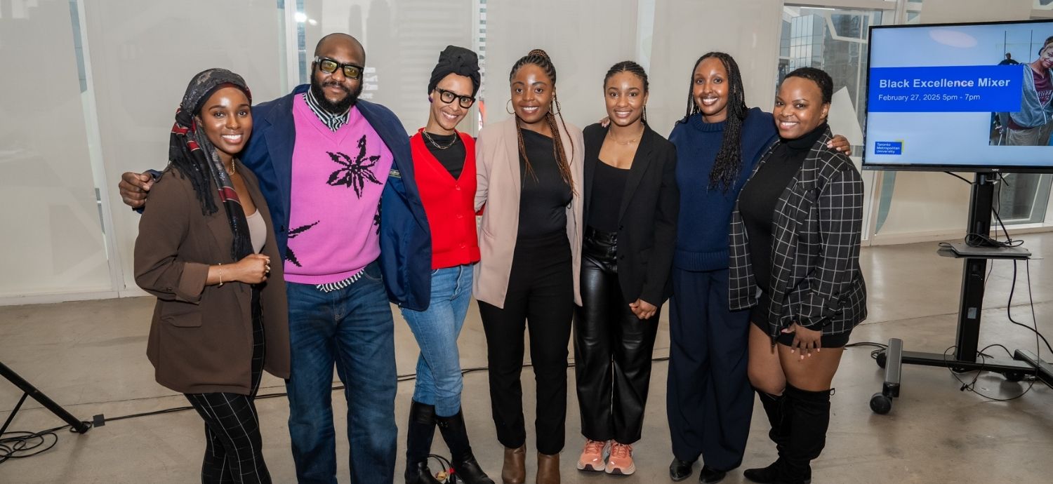 Seven people stand side by side and smile at a Black Excellence Mixer event, with an event screen visible in the background.