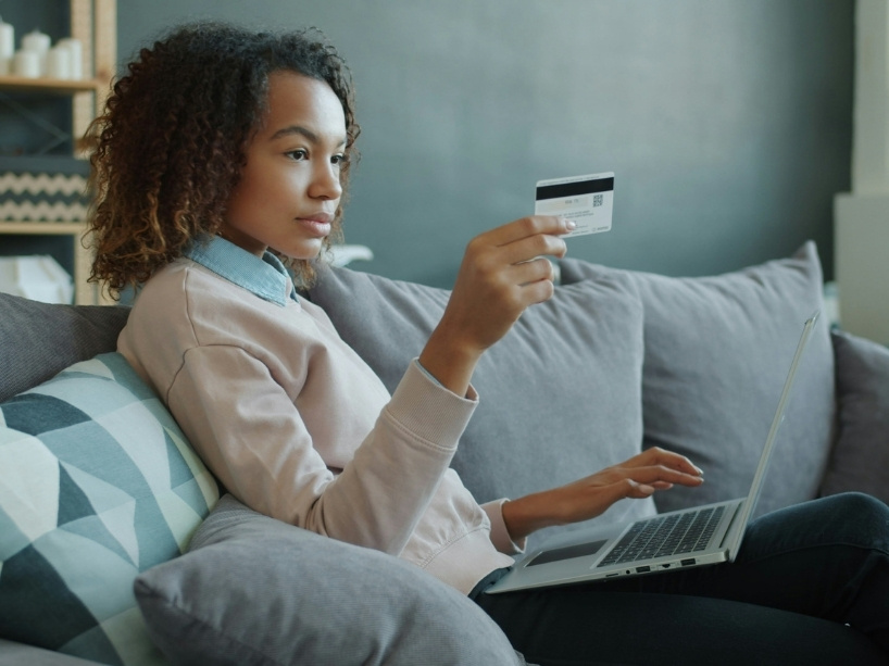 A woman sits on a couch with her laptop in front of her and credit card in hand.