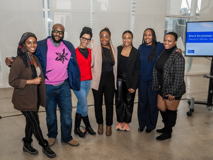 Seven people stand side by side and smile at a Black Excellence Mixer event, with an event screen visible in the background.
