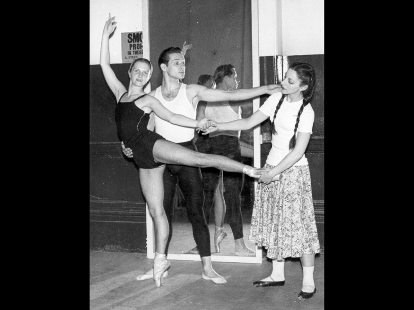 A female ballet dancer poses for the camera as another woman looks on and helps with her pose, and a male dancer poses behind her.