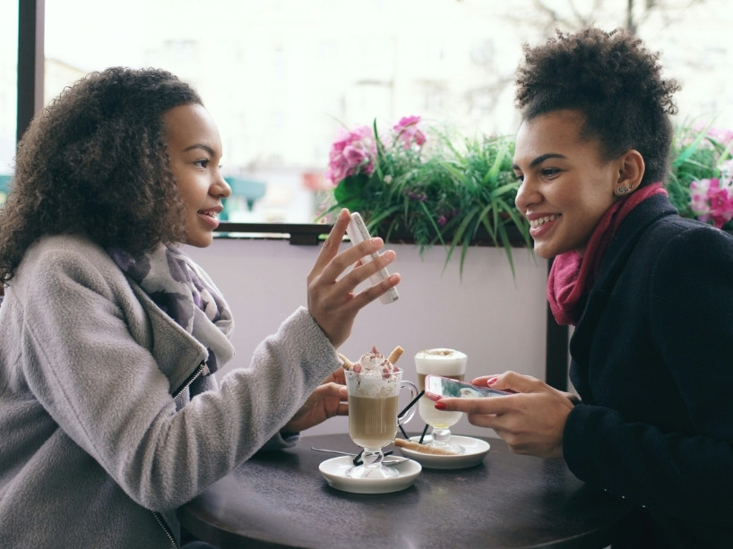 A woman reacts with a smile as another young woman shows her a cell phone screen.