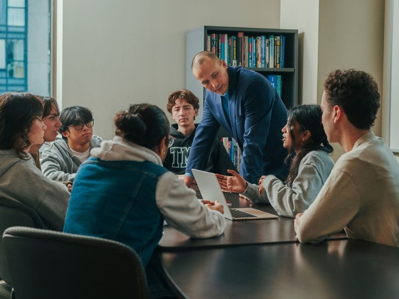 A professor among seven TMU students sitting at a table and listening to their peer who is chatting while looking at her open laptop.
