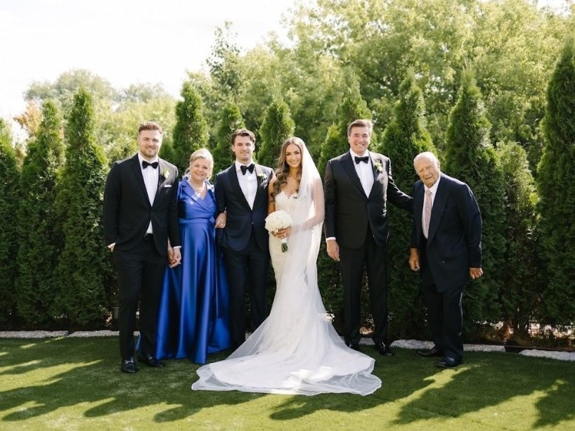 A bride and groom stand on the grass with family members in an area with trees and shrubs