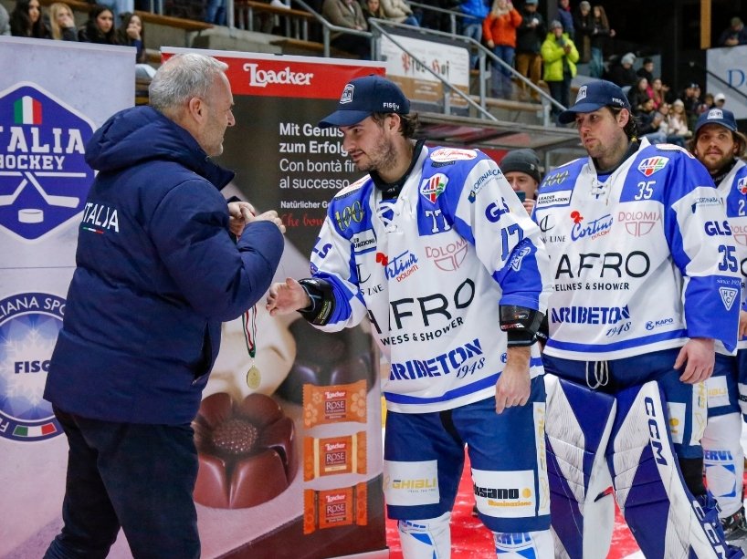A hockey player moves forward on the ice to receive a medal from a man in a jacket.