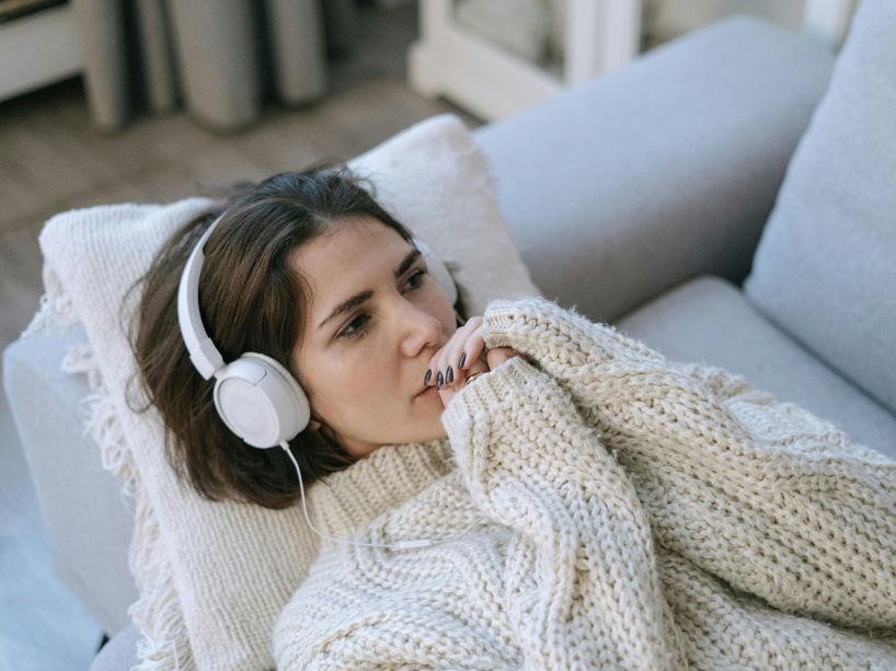 A young woman listens to music in her headphones while laying on a couch.