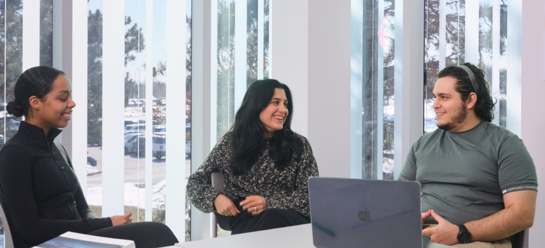 Two young women and a young man sit at a table by a window talking, with a laptop in front of them.